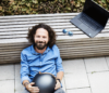 Foto eines mittelalten Mannes, der an einer hölzernen Sitzbank lehnt mit dem Laptop neben sich stehend und einen Mofahelm am Schoß hält